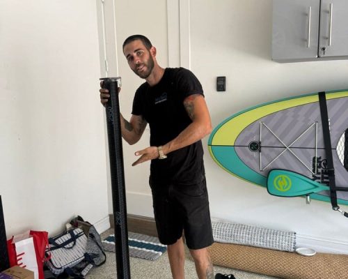 A man in black clothing stands in a garage, holding a long black cylindrical object and gesturing toward it. Behind him are a paddleboard and various boxes and shelves, suggesting he may be discussing Garage Door Repair or related repair services.